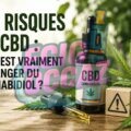 Illustration d’un flacon d’huile de CBD avec pipette posé sur une table en bois, entouré de feuilles de chanvre et d’un symbole d’avertissement, avec le titre « Les risques du CBD : Quel est vraiment le danger du cannabidiol ?